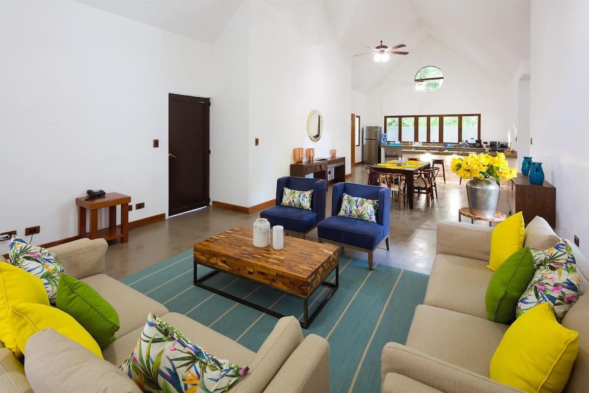 Uma sala espaçosa com um conjunto de sofás, uma mesa de centro e uma mesa de jantar com cadeiras. Uma cozinha aparece ao fundo, com uma geladeira e uma pia.