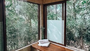 Une salle de bain avec une douche, un banc et une vue sur la verdure grâce à de grandes fenêtres.