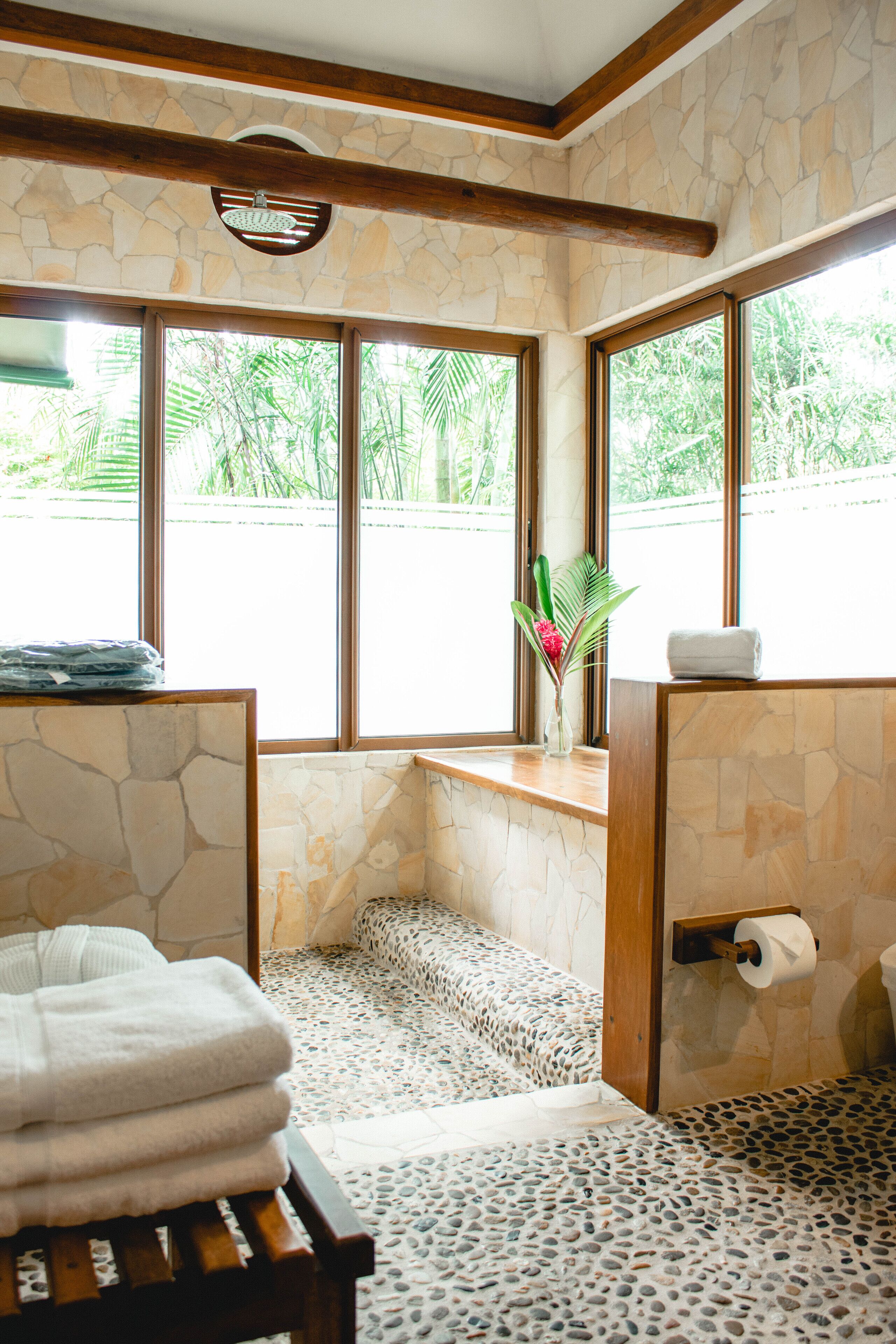 Un baño con paredes de piedra, un banco de madera, un perchero y un ventanal con vistas al verdor.