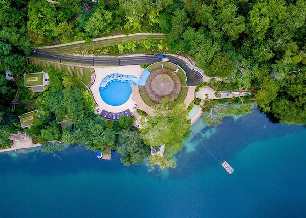 An aerial view of a resort with a central swimming pool, surrounded by lush greenery and a small boat docked near the water.