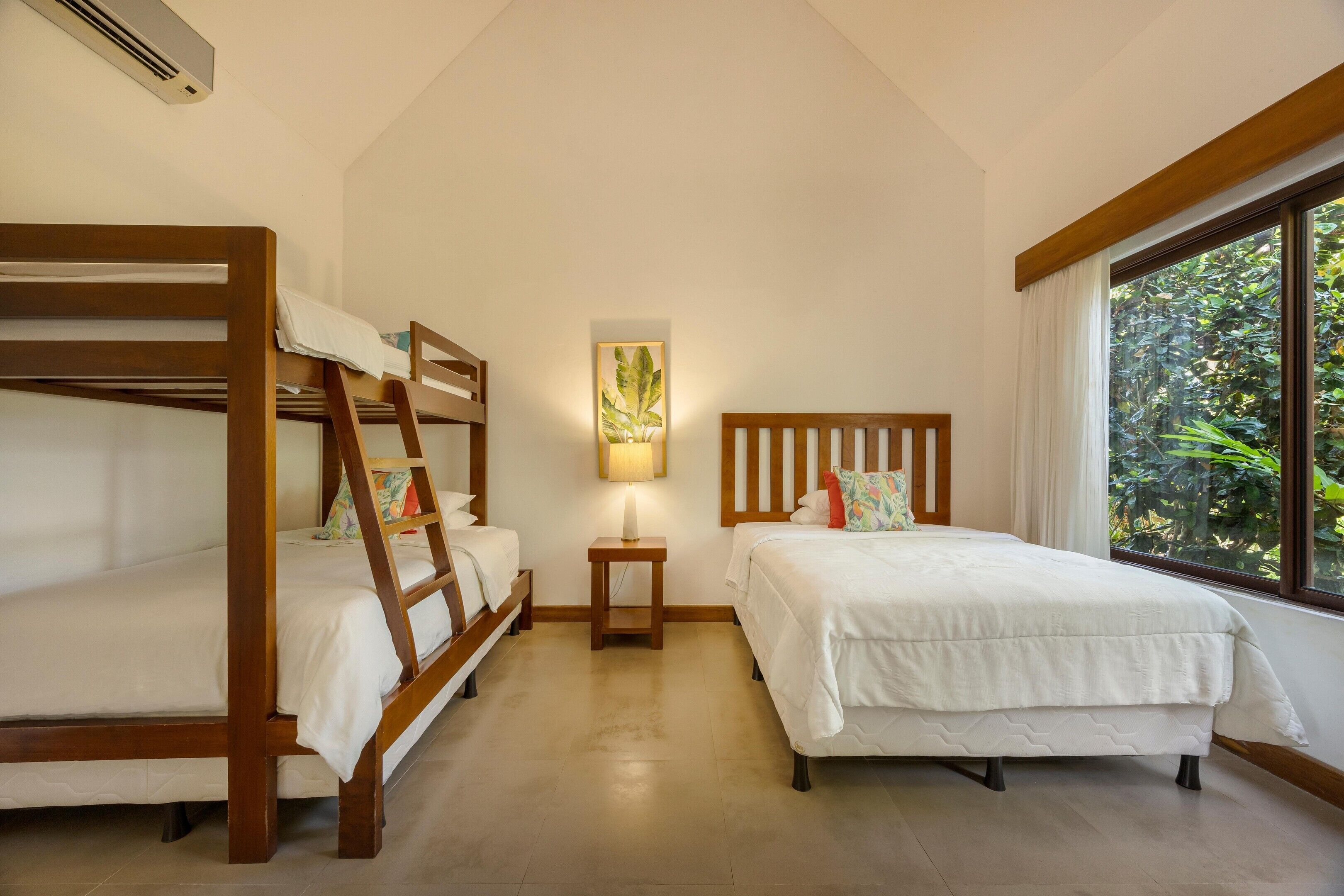 A room with bunk beds, a bedside table with a lamp, a wall-mounted artwork, and a window with a view of greenery.