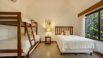 A room with bunk beds, a bedside table with a lamp, a wall-mounted artwork, and a window with a view of greenery.