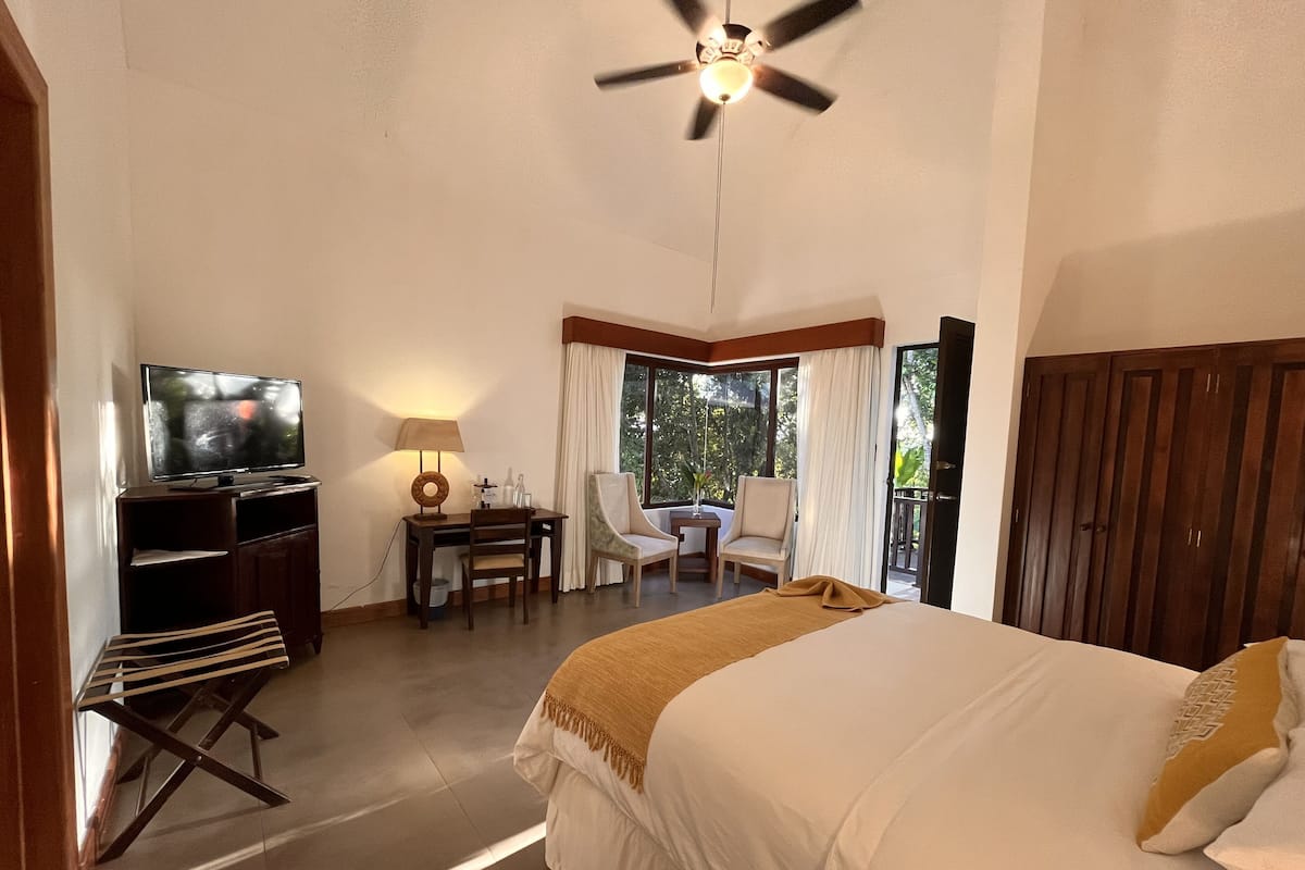 Quarto de hotel com cama, escrivaninha com luminária, televisão e varanda com cadeiras.