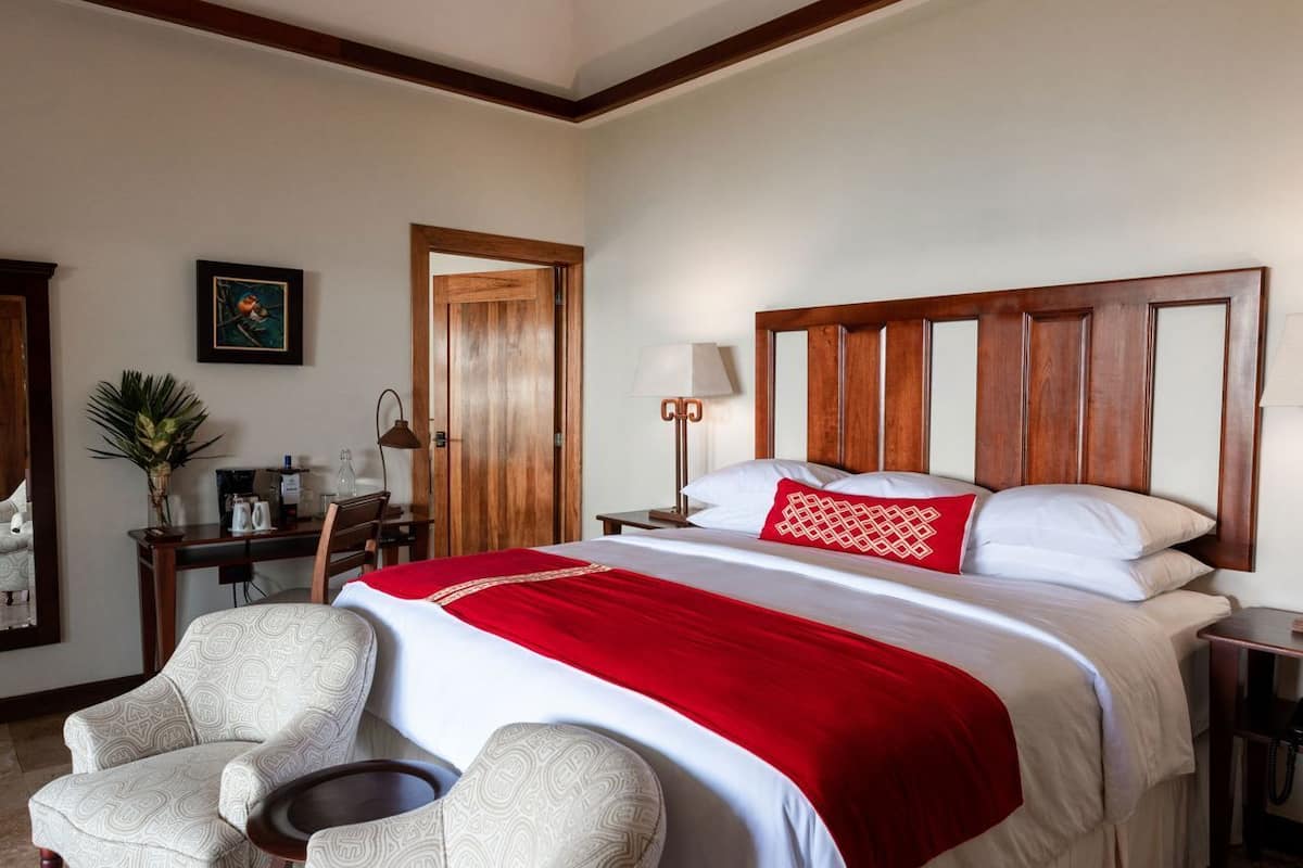 Quarto de hotel com uma cama grande, duas poltronas, uma escrivaninha e cabeceira de madeira.