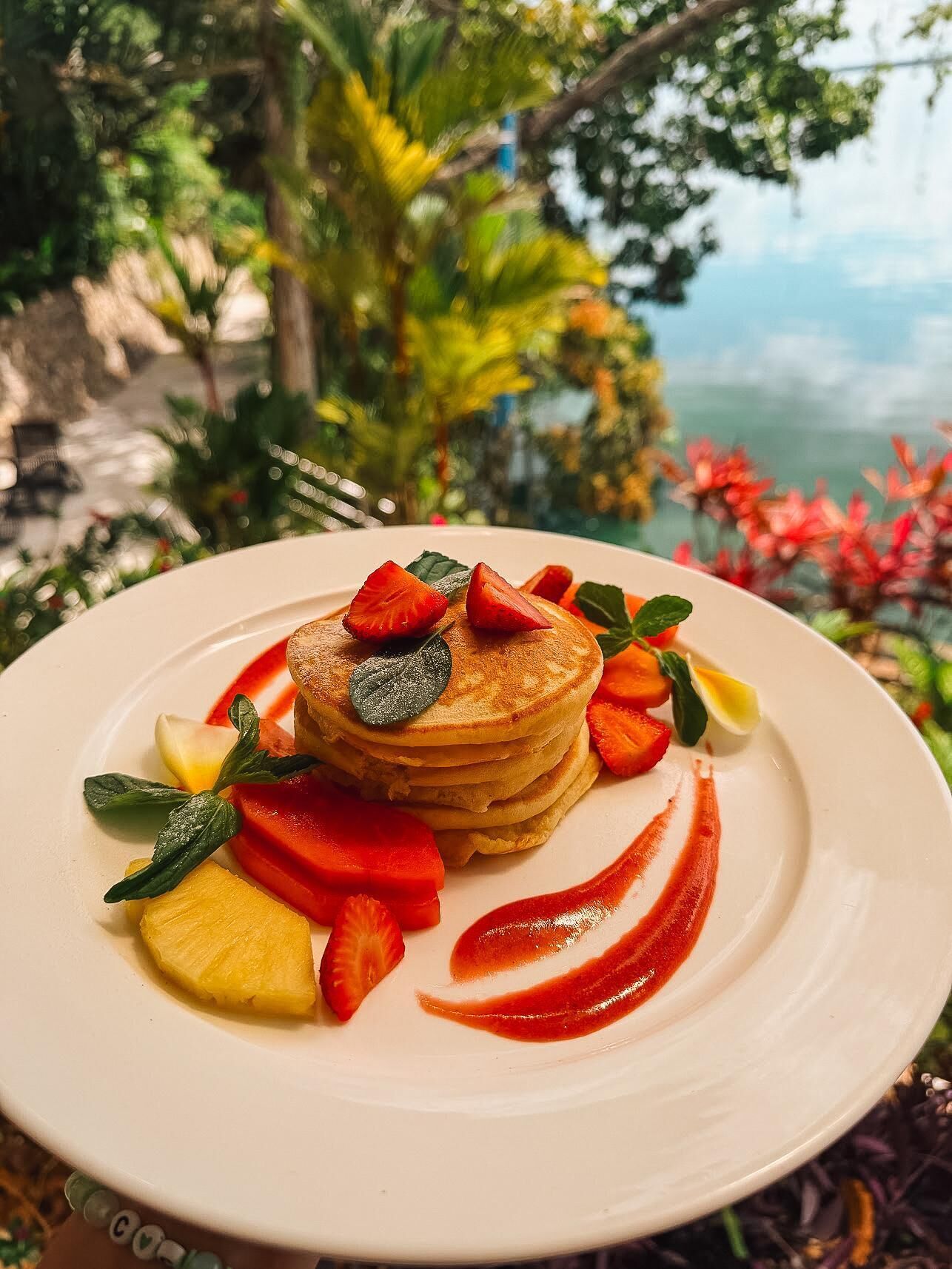 A stack of pancakes on a plate with strawberries, pineapple, mint leaves, and a red sauce.