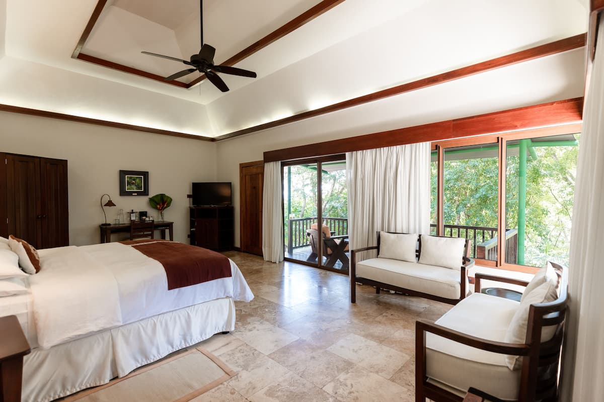 Um quarto espaçoso com uma cama grande, uma escrivaninha, televisão e varanda com vista para o verde.