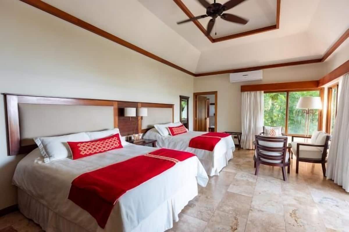 Quarto de hotel com três camas, ventilador de teto, escrivaninha com cadeira e janelas amplas.