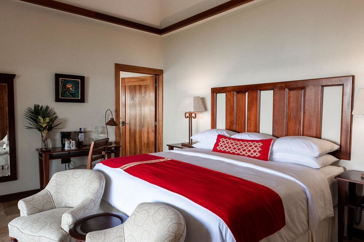 Une chambre d’hôtel avec un grand lit, deux fauteuils, une petite table, un bureau avec une lampe et une porte.