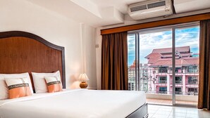 A hotel room with a large bed, a wooden headboard, a window with curtains, and a view of buildings outside. - Piyada Residence by Nexottel (Pattaya)