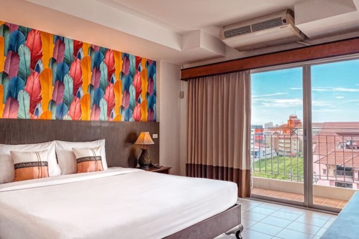 A hotel room with a large bed, a colorful wall mural, a window with curtains, and an air conditioning unit.