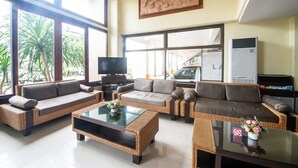 A spacious living area with wicker furniture, a flat-screen TV, and large windows offering a view of palm trees. - Piyada Residence by Nexottel (Pattaya)
