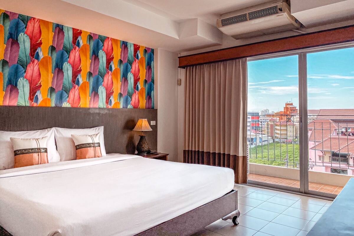 Une chambre d’hôtel avec un grand lit, un mur décoré d’un motif coloré représentant des plumes, une table de chevet avec une lampe et une grande fenêtre donnant sur la ville.