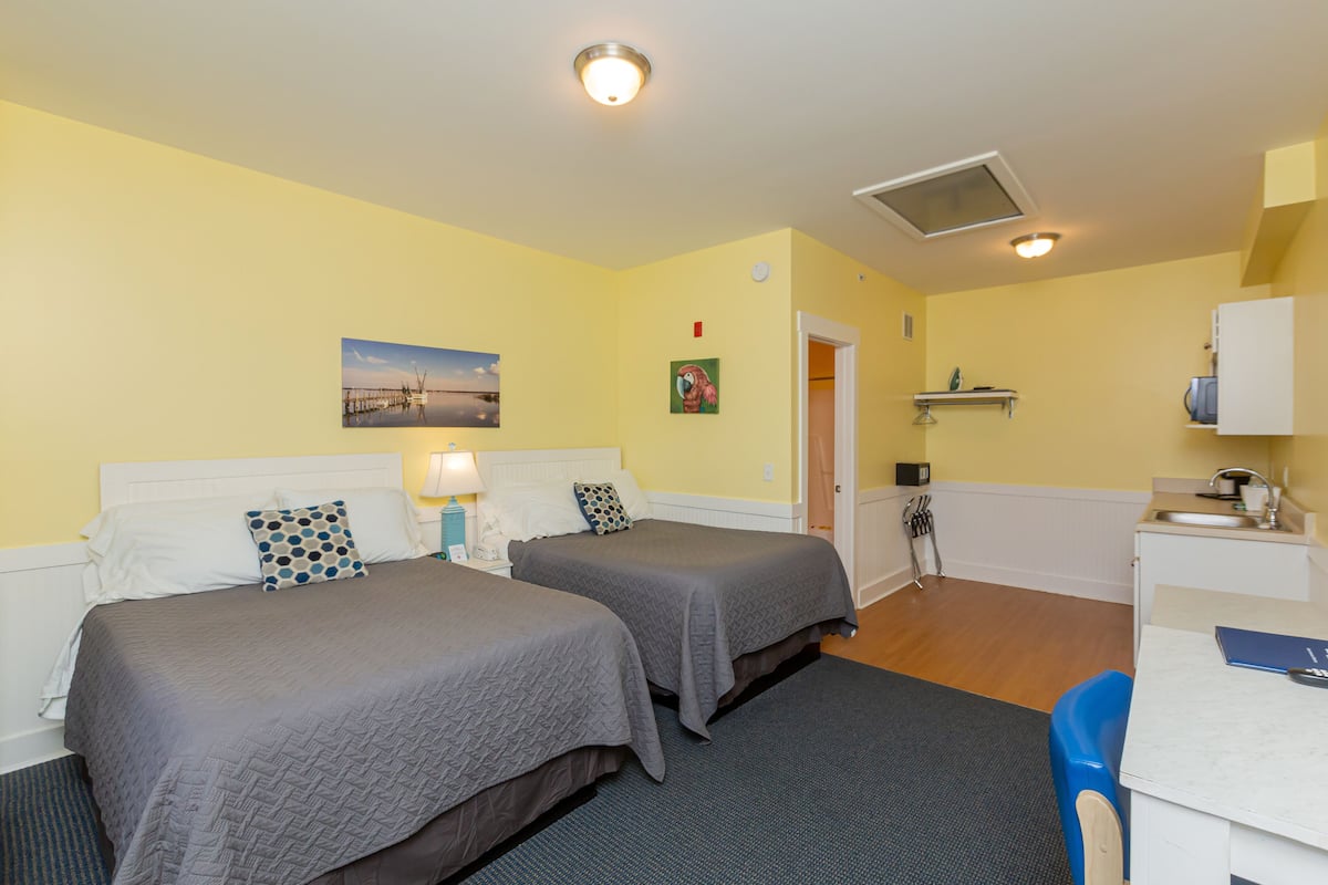 A hotel room with two beds, a desk, a chair, a sink, and a small shelf.