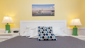 A hotel room with a bed, two nightstands, two lamps, a painting, and a patterned pillow. - Topsail Shores Inn (Sneads Ferry)