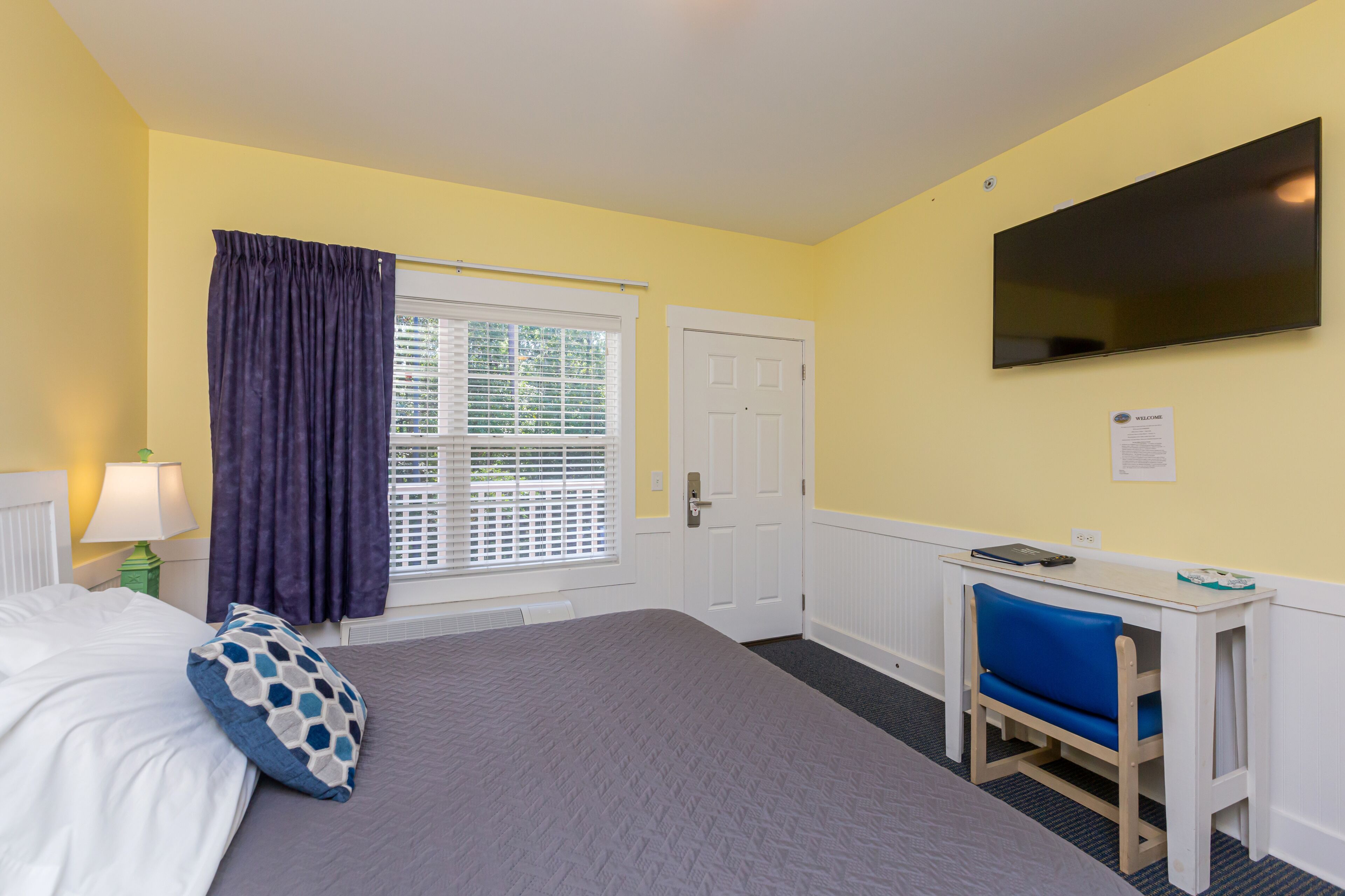 A hotel room with a bed, a desk with a chair, a TV, and a window with curtains.