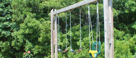 A wooden swing set with four swings, a green picnic table, and a wooded background.