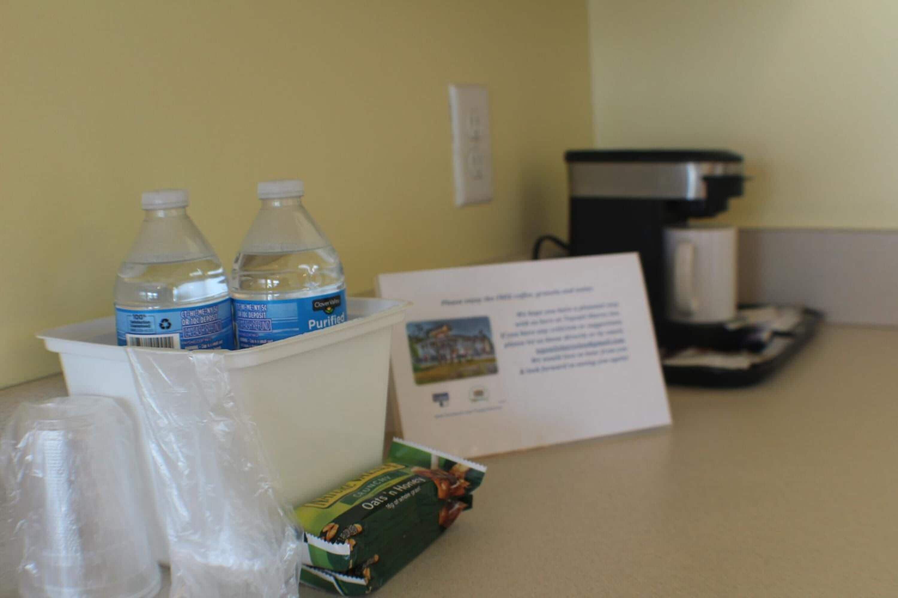 A hotel room counter with bottled water, a coffee maker, a menu, and a small box of snacks.