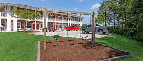 A playground with swings and a sandbox, a building with multiple stories and red doors, and cars parked in front.