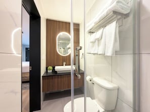 Un baño moderno con cabina de ducha de vidrio, inodoro blanco, tocador de madera con espejo y encimera negra.