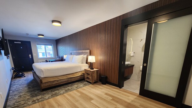 Una habitación de hotel moderna con una cama de madera, una mesita de noche, una lámpara, un baño con puerta de vidrio y vistas al exterior.