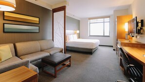 A hotel room with a sofa, a bed, a desk with a chair, a TV, and a large window.