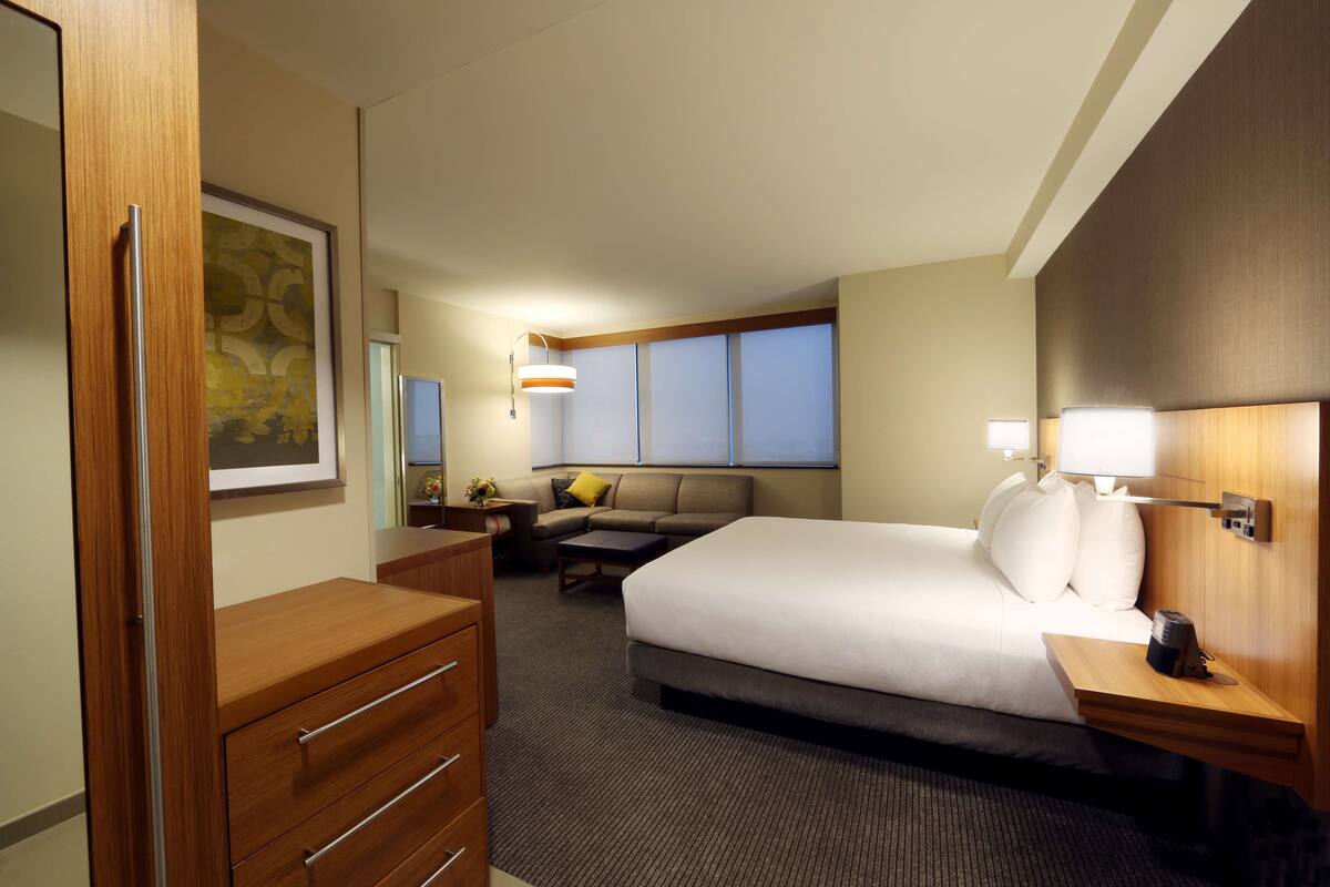 Ein Hotelzimmer mit Bett, Schreibtisch, Kommode, Sofa und einem Fenster mit Jalousien.