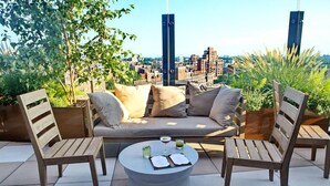 A rooftop terrace with a sofa, two wooden chairs, a round table with a menu, and a city view.