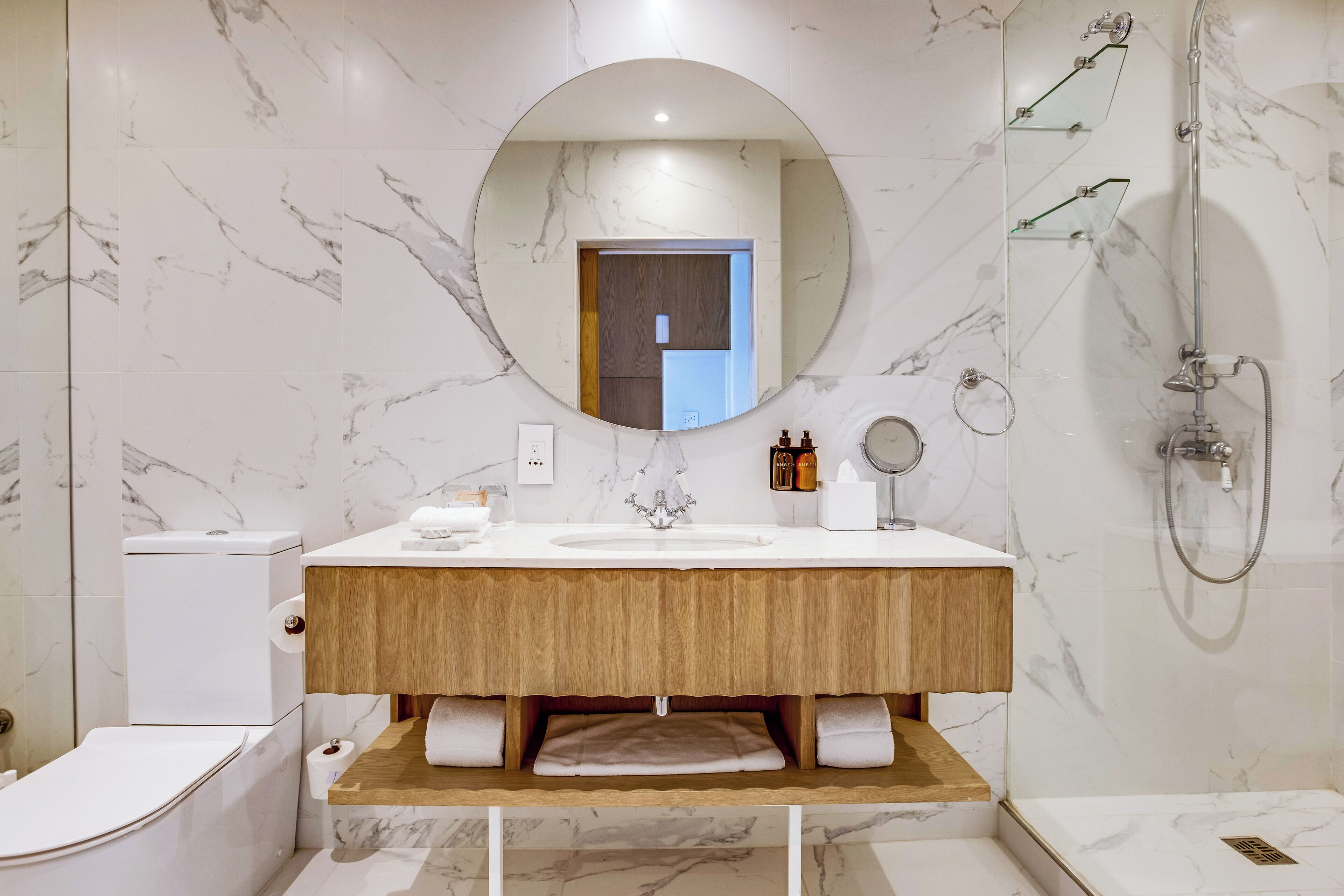A modern bathroom with a marble backsplash, a wooden vanity, a round mirror, a toilet, and a shower.