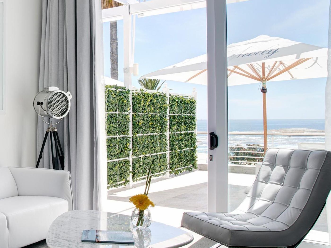 A modern living room with a white sofa, a grey armchair, a glass coffee table, and a floor lamp. There is a large window with a view of the ocean and a white umbrella.
