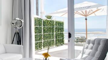 A modern living room with a white sofa, a grey armchair, a glass coffee table, and a floor lamp. There is a large window with a view of the ocean and a white umbrella.