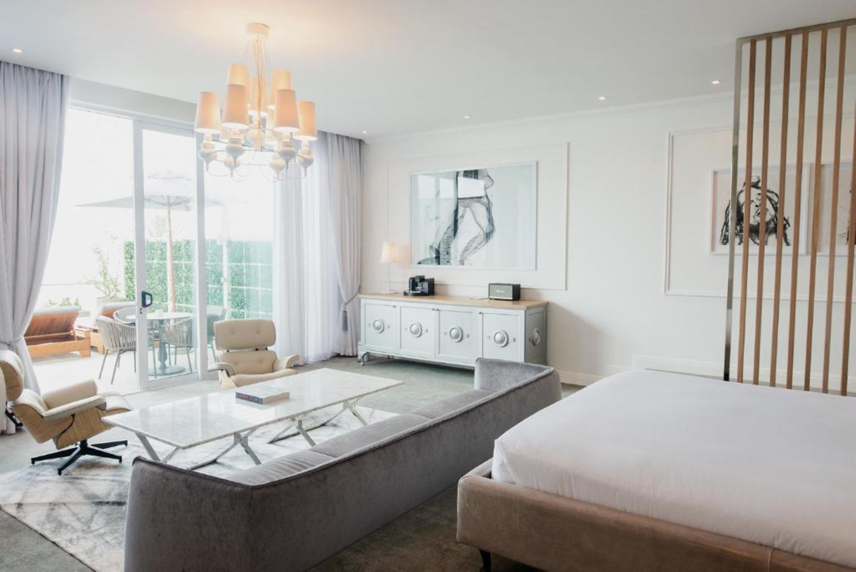 Una habitación de hotel moderna con una cama grande, un sofá gris, una mesa de centro de vidrio, un aparador blanco y un amplio ventanal con cortinas.
