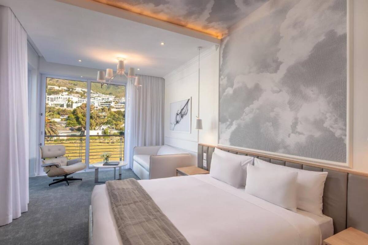 Une chambre d’hôtel dotée d’un grand lit, d’un coin salon avec un fauteuil et une petite table, offrant une vue sur le paysage urbain grâce à de grandes fenêtres, et ornée d’une fresque murale sur le thème des nuages.