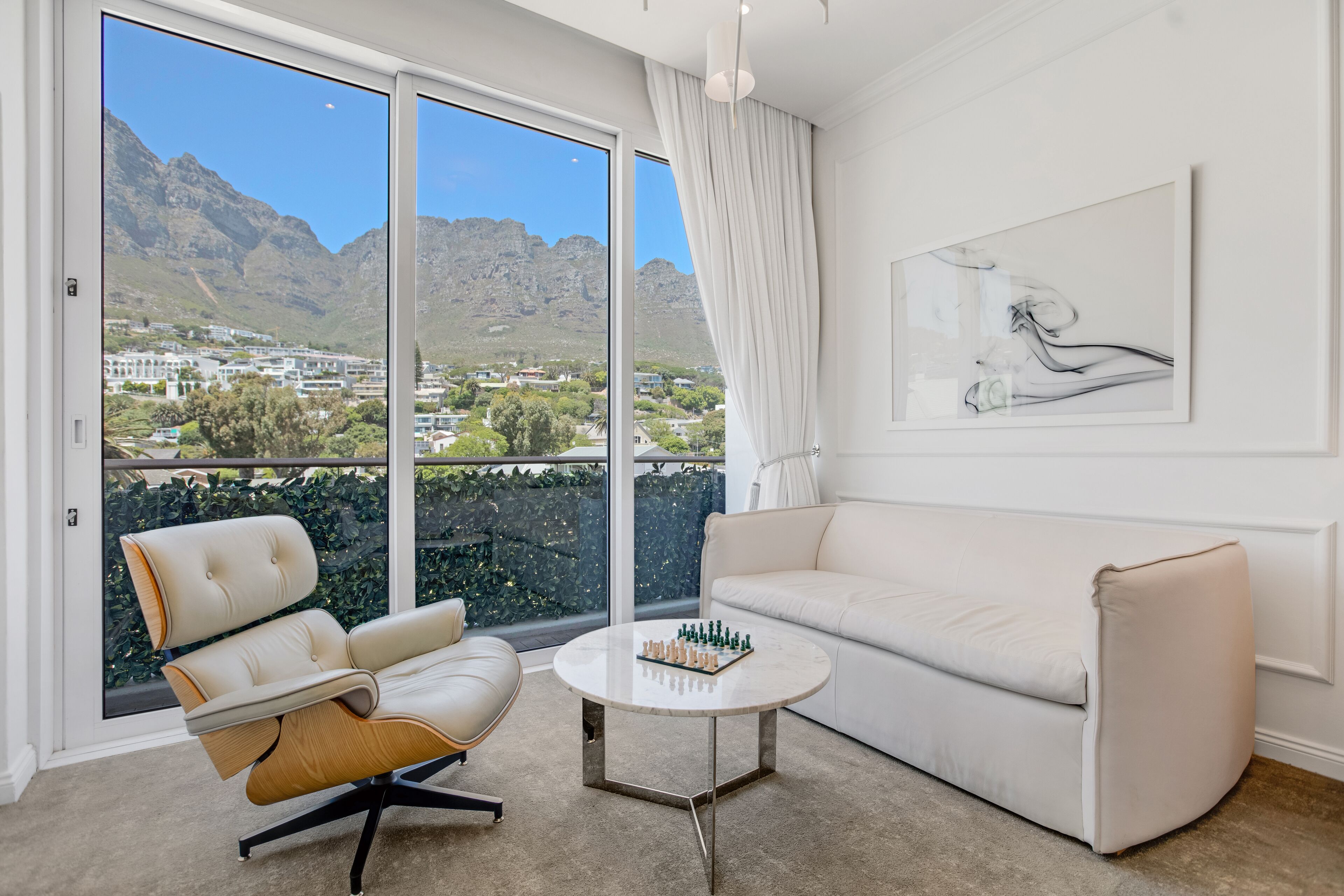 Una sala de estar moderna con vistas a las montañas, un sofá blanco, un sillón de cuero y un tablero de ajedrez sobre una mesa de centro.