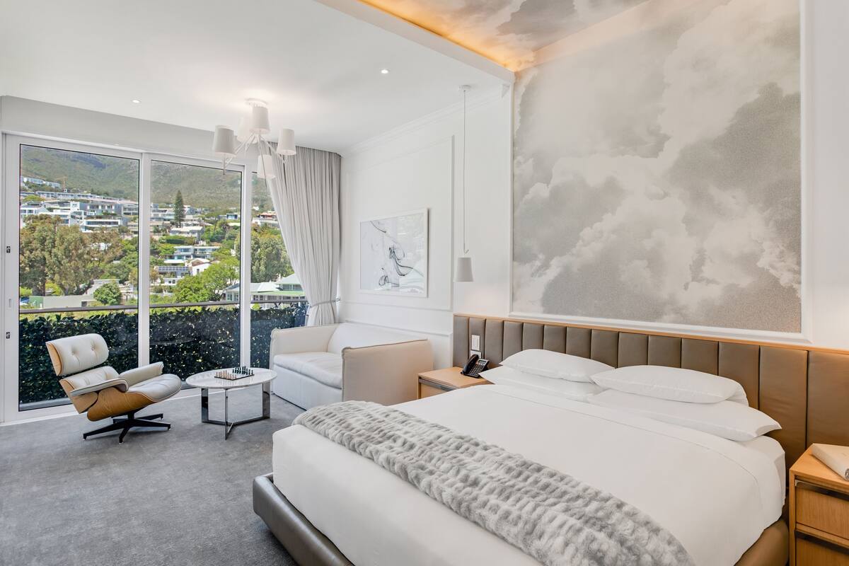 Une chambre d’hôtel moderne, dotée d’un grand lit, d’un canapé, d’un fauteuil, d’une petite table et offrant une vue sur le paysage urbain grâce à de grandes fenêtres.
