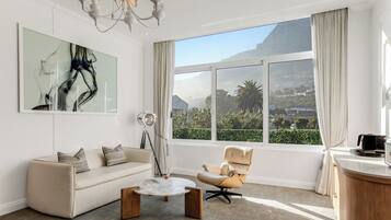 A modern living room with a sofa, armchair, and coffee table, a large abstract painting, and a view of a mountainous landscape through a large window.