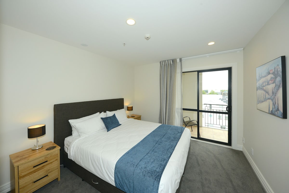 A bedroom with a bed, bedside tables, a nightstand, a balcony with a view, and a painting on the wall.