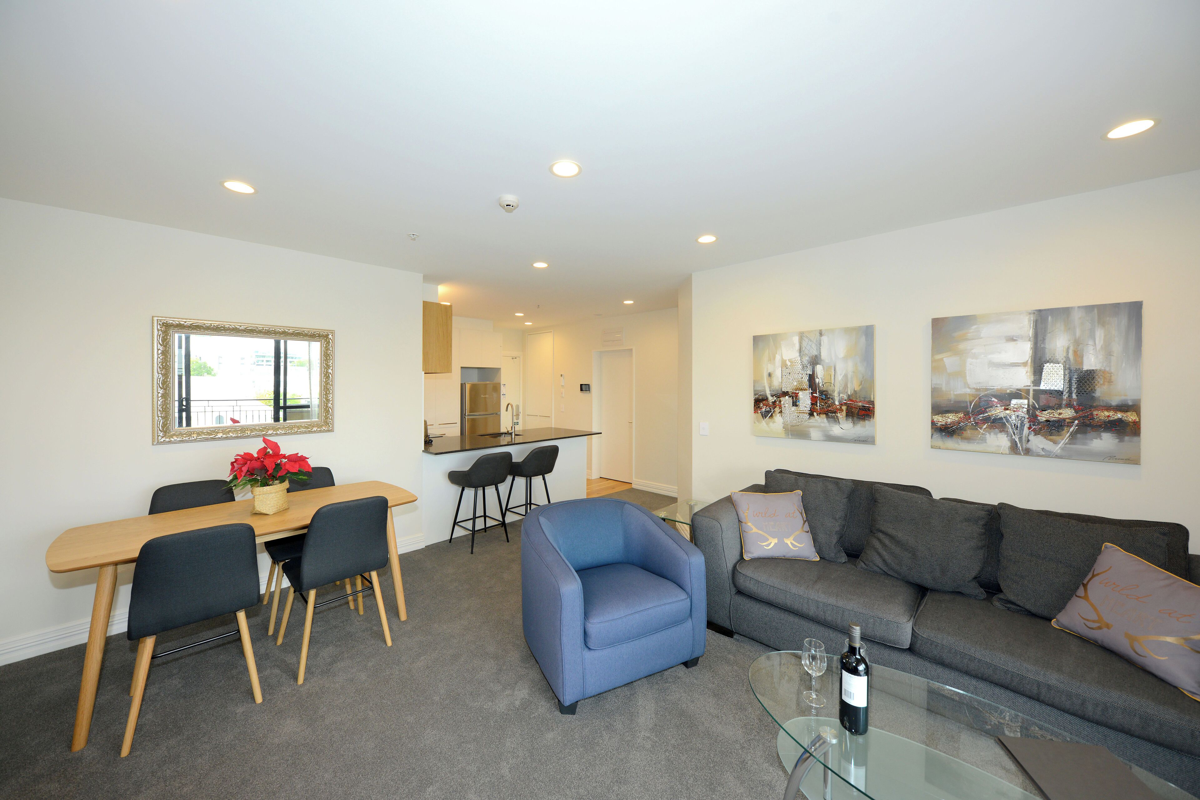 A modern living room with a dining area, grey sofa, and a glass coffee table with a bottle of wine.