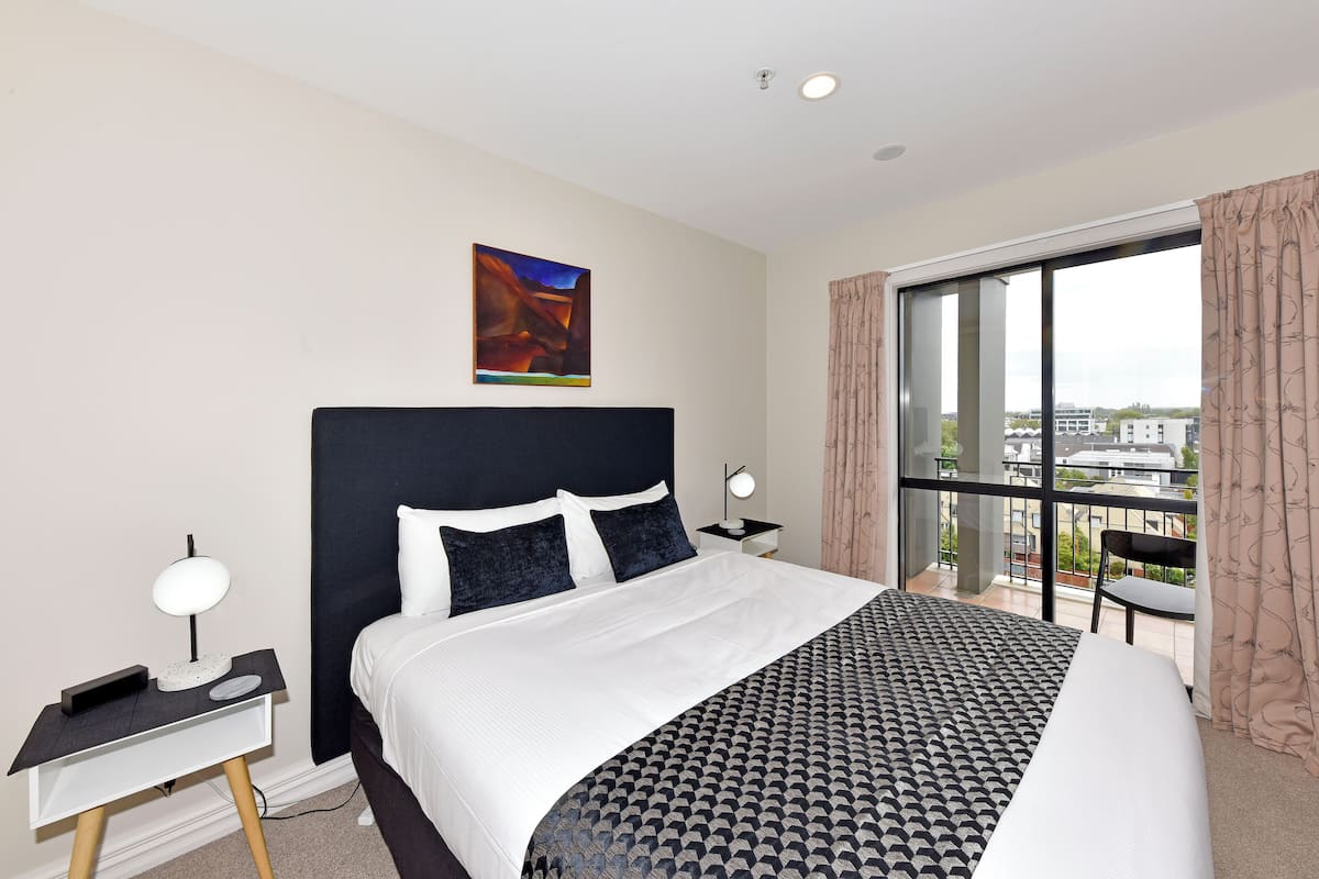 A modern hotel room with a large bed, a desk, a chair, and a view of the city through large windows.