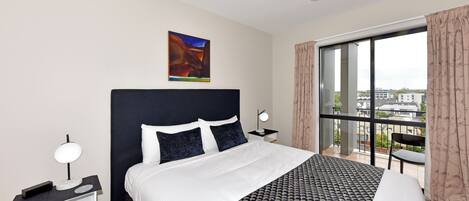 A modern hotel room with a large bed, a desk, a chair, and a view of the city through large windows.