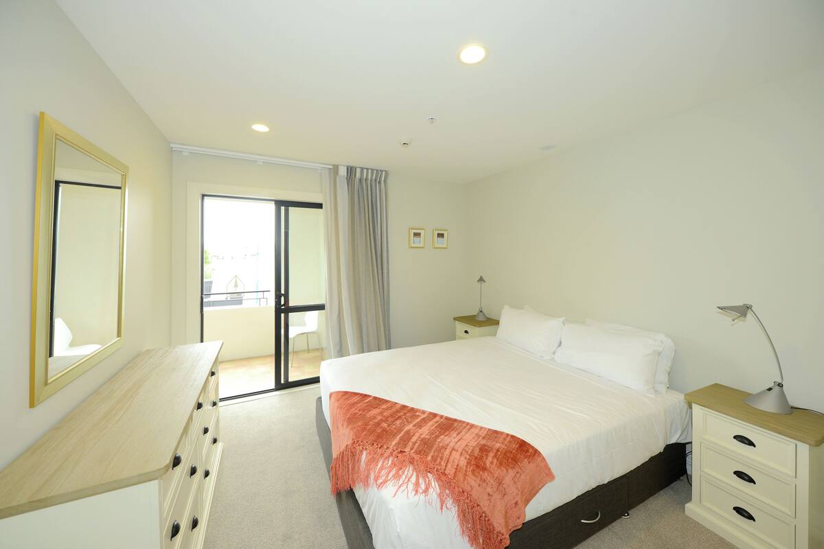 A bedroom with a bed, a dresser, a nightstand, a mirror, and a window with curtains.