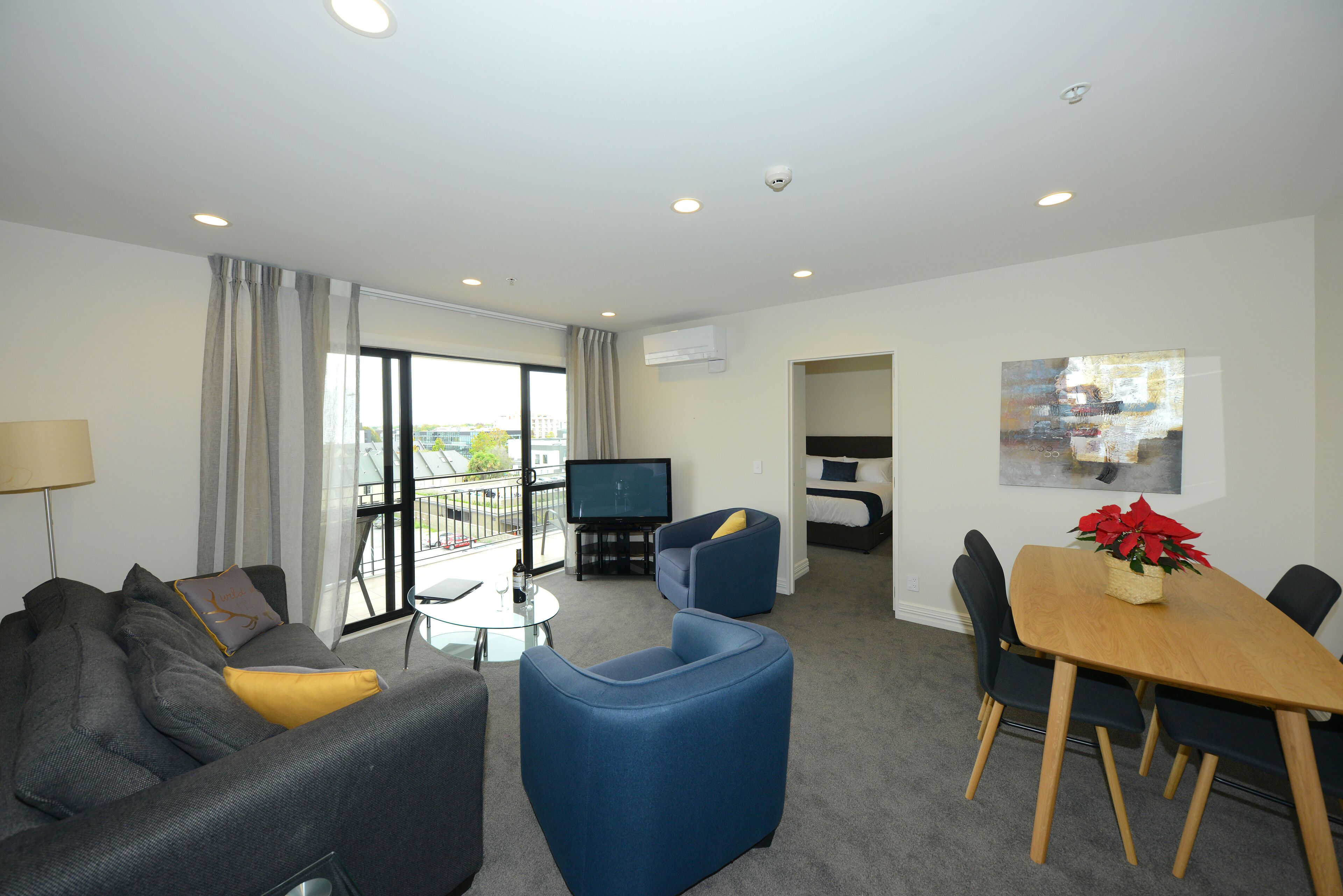 A modern living room with a grey sofa, blue armchair, and a wooden dining table with chairs. A flat-screen TV is mounted on the wall, and there is a balcony visible through the large window.