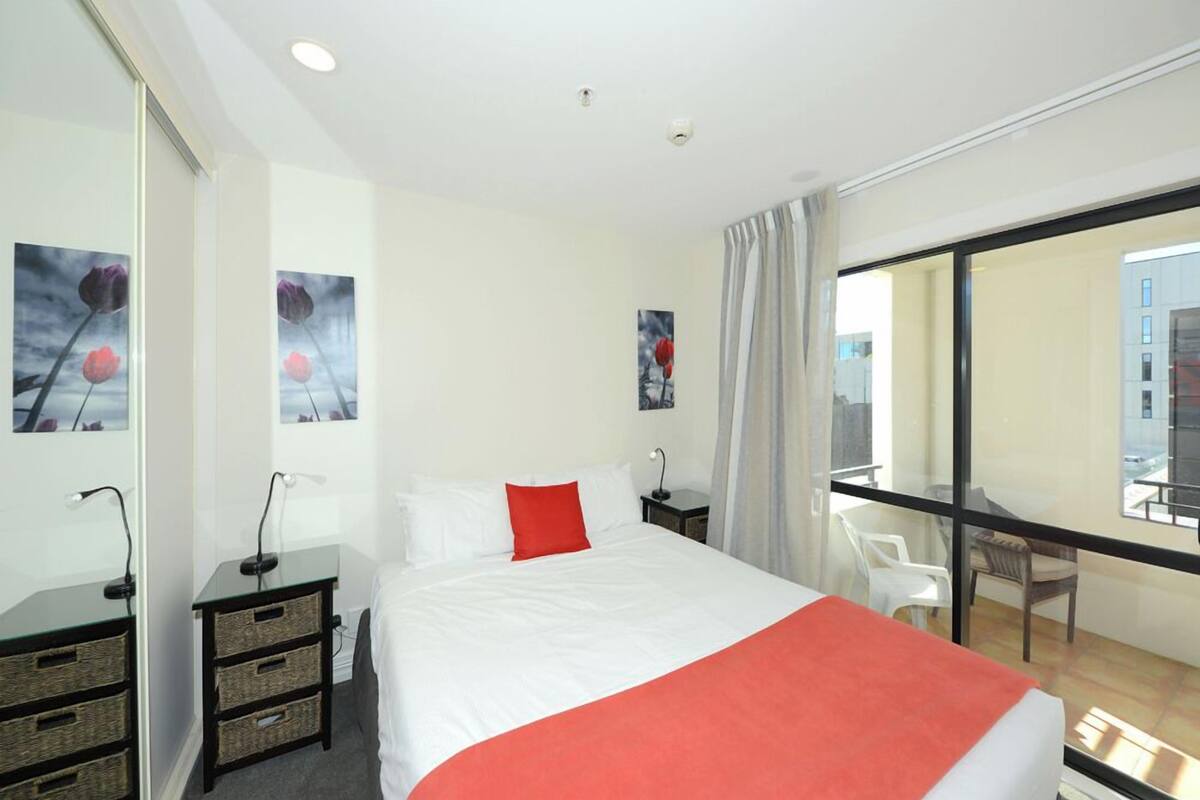 A hotel room with a large bed, a desk with a lamp, a chair, and a balcony with a view of buildings.