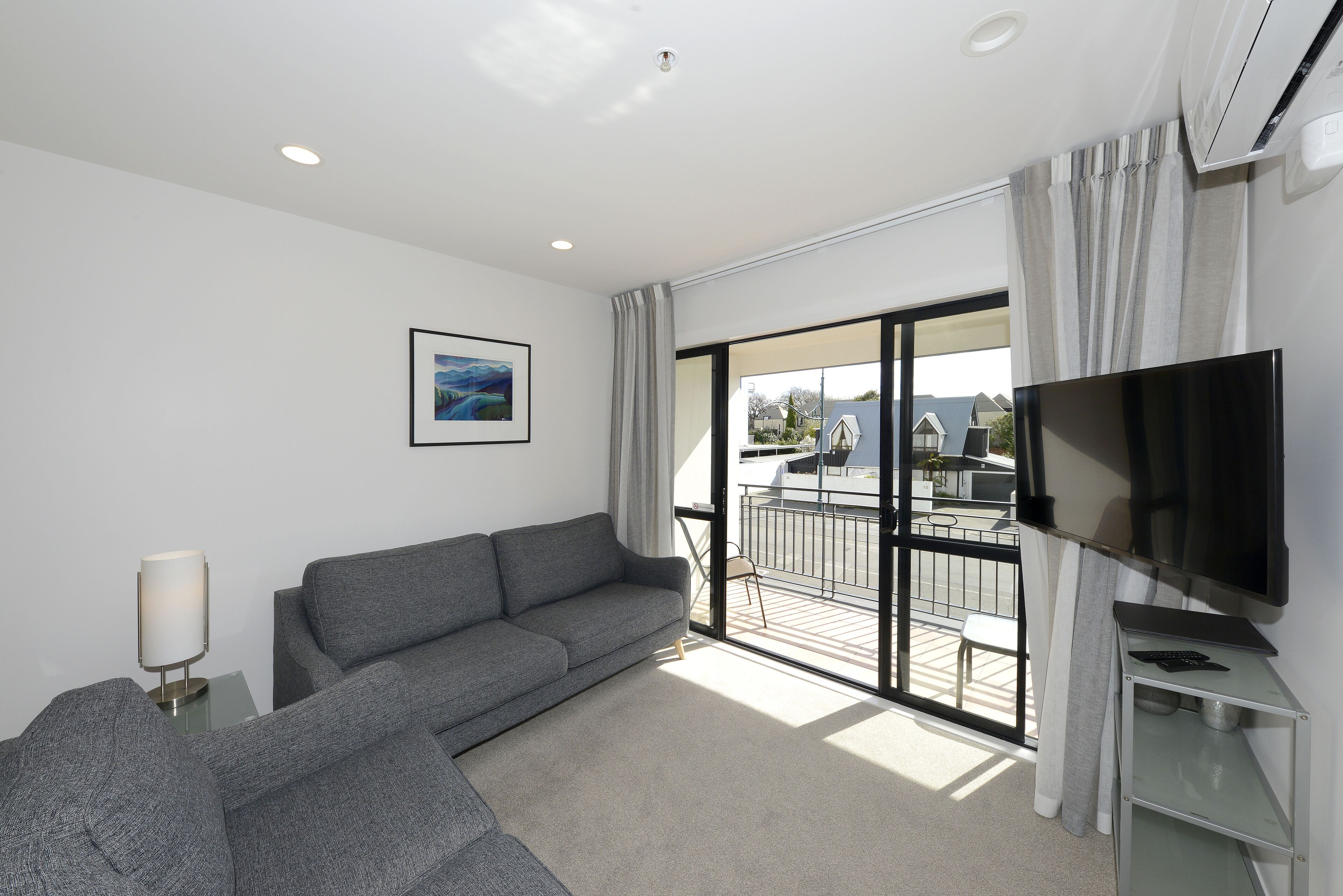 A modern living room with a grey sofa, a flat-screen TV, and a large window with a view of a balcony.