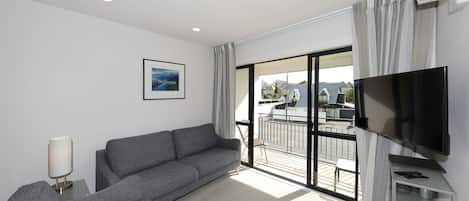 A modern living room with a grey sofa, a flat-screen TV, and a large window with a view of a balcony.