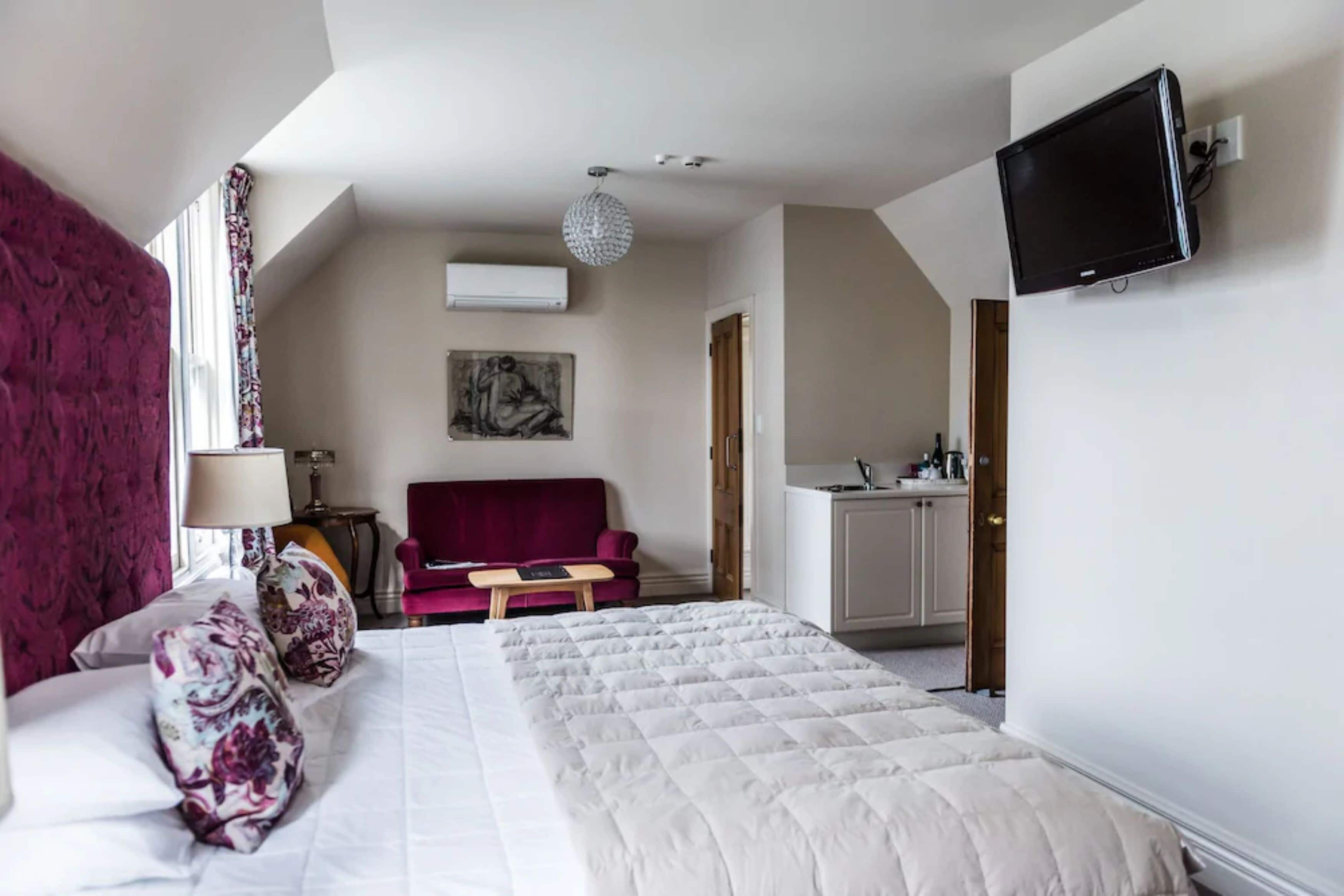 Quarto de hotel com uma cama, um sofá vermelho, uma mesinha, uma TV na parede e uma cozinha compacta.