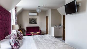 Quarto de hotel com uma cama, um sofá vermelho, uma mesinha, uma TV na parede e uma cozinha compacta.