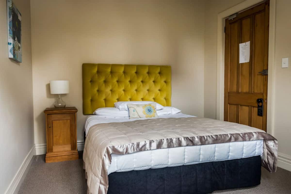 Un dormitorio con cabecera amarilla acolchada, cama con colcha beige y oscura, mesita de noche de madera con lámpara, y una puerta de madera con un cartel.