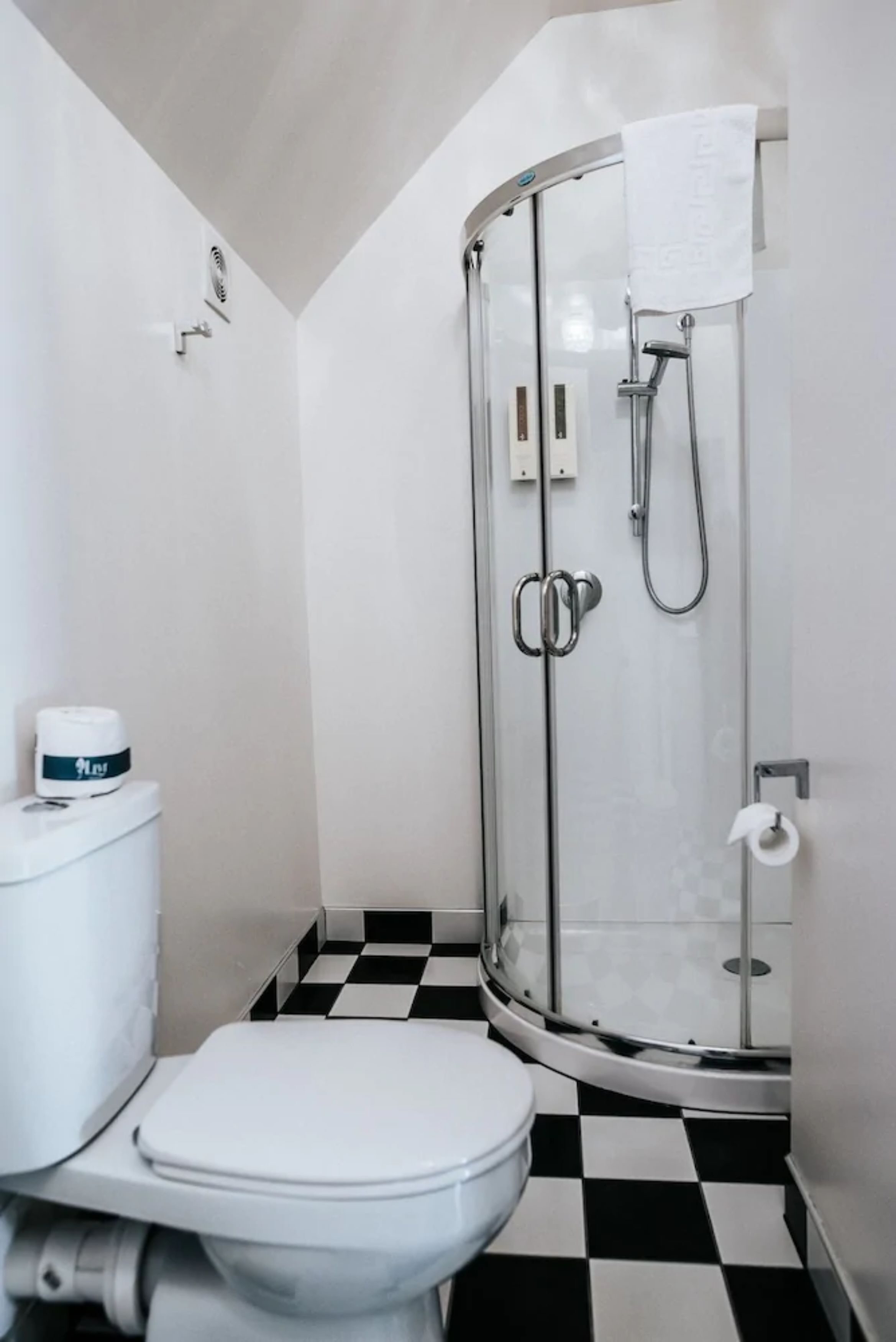Un baño con inodoro blanco, cabina de ducha de vidrio y suelo a cuadros blanco y negro.
