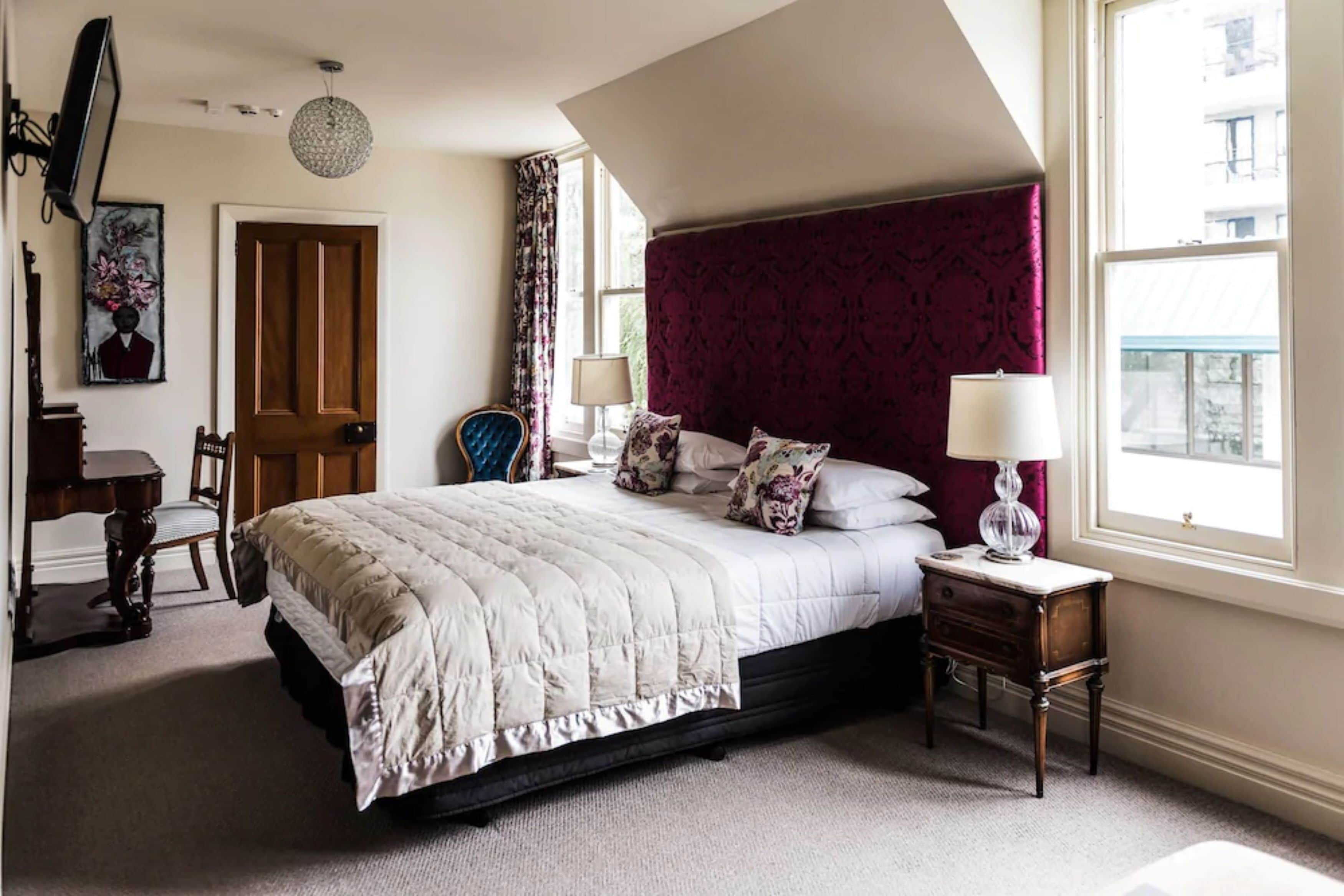 A bedroom with a large bed, a nightstand, a chair, a desk, and a window with curtains.