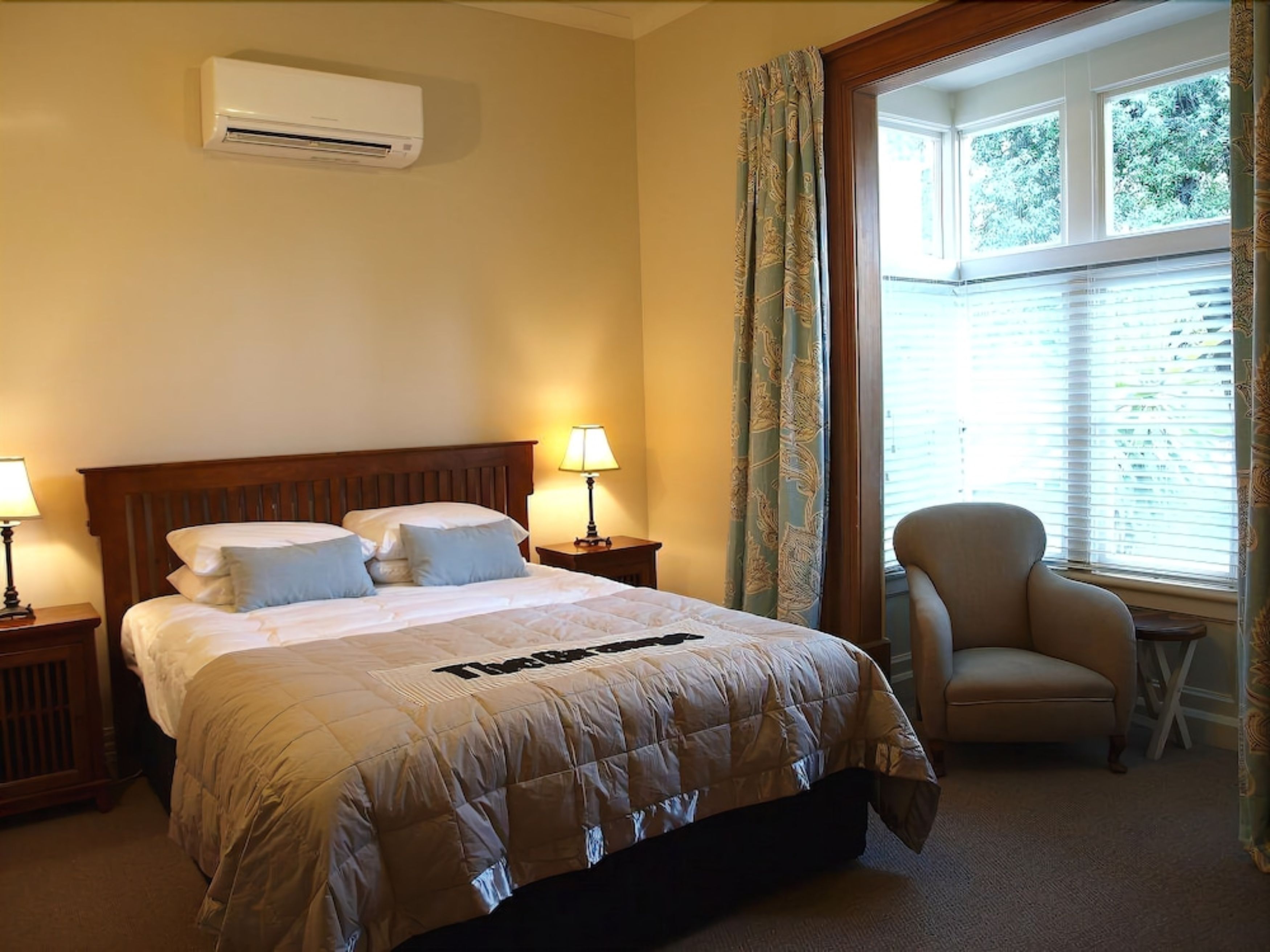Habitación de hotel con cama, dos mesitas de noche con lámparas, una silla y una ventana con persianas.